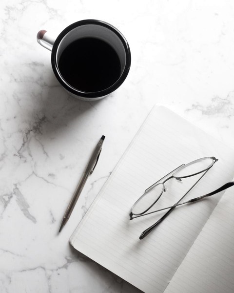 Reading glasses resting on an open notebook beside a pen and a cup of coffee